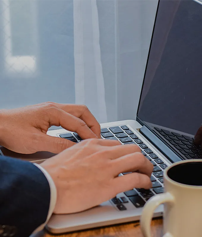 デスクでノートパソコンのキーボードを叩く手元、パソコンの横にはコーヒーカップが置かれている